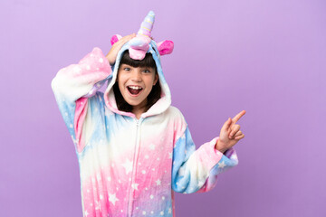Little kid wearing a unicorn pajama isolated on purple background surprised and pointing finger to the side © luismolinero