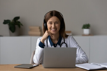 Head shot portrait smiling female doctor therapist in wireless headphones sitting at work desk with laptop, professional practitioner gp looking at camera, consulting patient online by video call