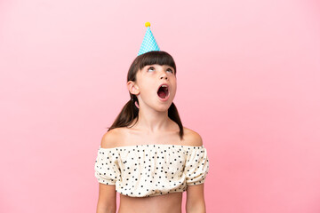 Little caucasian kid with birthday hat isolated on pink background looking up and with surprised...