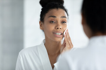 Head shot happy attractive millennial african american woman in bathrobe looking in mirror applying toner with cotton pad on face after bath, preparing face for daily nude makeup, skincare concept. - Powered by Adobe