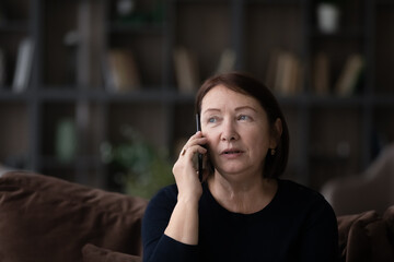Head shot close up of serious mature woman talking on phone at home sitting on couch, senior female...