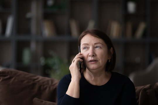 Head shot close up of serious mature woman talking on phone at home sitting on couch, senior female holding mobile device smartphone, chatting with relatives or friends, making or answering call