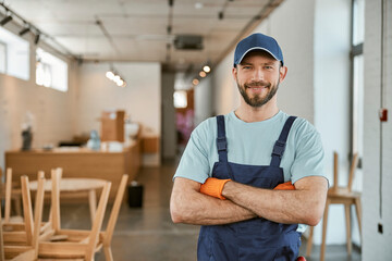 Joyful male worker in cap keeping arms crossed - obrazy, fototapety, plakaty