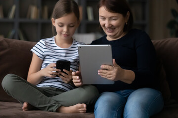 Happy mature grandmother with granddaughter using modern devices together, sitting on couch at home, little girl and senior woman using smartphone and tablet, spending leisure time with gadgets