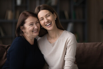 Happy mature woman with grownup daughter hugging sitting on couch, smiling young woman and senior...