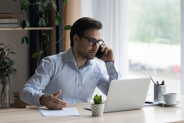 Focused smiling young businessman in glasses consulting client by phone call, working on computer in modern office. Smart millennial male manager employee multitasking, negotiating project distantly.