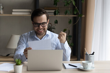 Happy sincere young man in glasses making yes gesture looking at document, reading amazing news in paper correspondence, getting bank loan approval, dream job offer, celebrating signing contract deal.