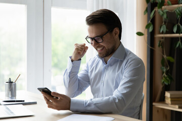 Happy emotional young man in eyewear looking at cellphone screen, celebrating online lottery...