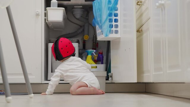 Toddler wearing safety helmet opening cabinet in kitchen child playing with trash can, baby safety proofing at home. High quality 4k footage