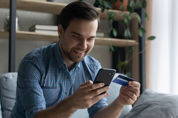 Happy young man using mobile shopping application, entering payment information from credit bank card, purchasing goods or services online, satisfied with fast money transfer, cyber security.