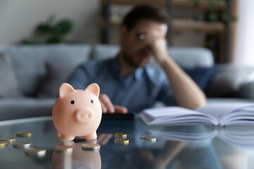 Close up focus on piggybank on table, young man feeling stressed calculating expenditures or taxes,...
