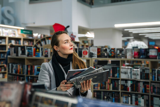 A Beautiful Young Woman Chooses A Comic Book For A Gift In A Bookstore To Buy It. Stands In Front Of A Shelf Of Books And Holds A Stack Of Comics Comparing Them.