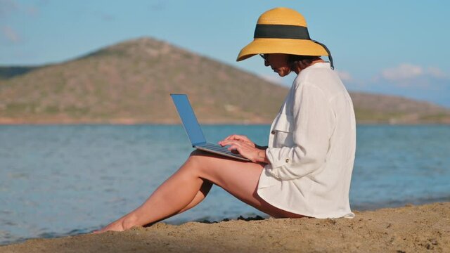 Mature woman in hat using laptop on sea beach