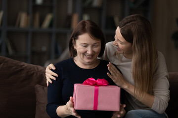 Loving grown-up daughter congratulating happy mature mother, hugging touching shoulders, presenting pink gift box, sitting on couch, family celebrating birthday or mothers day at home together