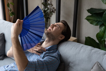 Exhausted young man waving paper fan, breathing fresh cooled air, suffering from high temperature...