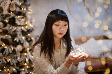 A young, beautiful Korean Asian woman holds a lighted festive garland in her cozy home with dreams of positive emotions near the Christmas tree in the evening. The concept of New Year and Christmas.