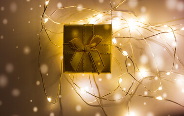 Yellow gift box with bow lies on table amidst glowing tangled led garland lights flatly. New Year 2022, Christmas. Magical winter evening. Gold box with present inside. Falling snowflakes background.