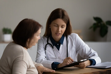 Female doctor therapist consulting mature woman at appointment, holding clipboard, explaining medical checkup results giving recommendations to middle aged patient, discussing insurance contract