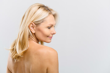 Charming good-looking blonde middle-aged woman with naked shoulders, looks down and smiling over grey background. Anti-aging treatment and care. Spa and relax, cosmetology procedure