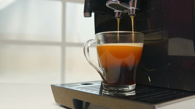 Coffee from machine is poured into cup at home or in office close-up. Female hand takes glass with Americano made by automatic coffee machine. Concept of morning begins with cup of aromatic coffee dri
