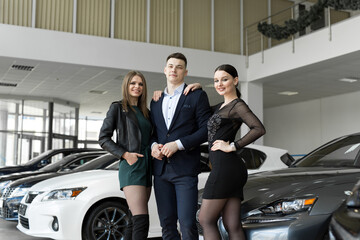 Friends two women and a man embrace against the background of new cars in a car dealership.