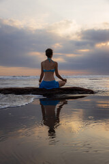 Fototapeta premium Sunset yoga. Caucasian woman sitting on the stone in Lotus pose. Padmasana. Hands in gyan mudra. Beach in Bali. View from back. Copy space. Yoga retreat. Water reflection. Healthy life. Zen life.