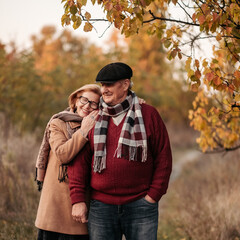 Elderly man lovingly looks at his elegant pensioner, hugs and enjoys her beauty, senior married couple dancing in autumn park, celebrating anniversary, affection
