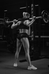 Naklejka na ściany i meble A photo from behind of a fit woman who is squatting with a barbell in the gym