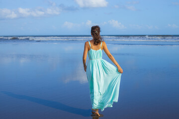 Young woman walking barefoot on empty beach. Full body portrait. Slim Caucasian woman wearing long dress. View from back. Summer sunlight. Blue sky. Travel concept. Bali, Indonesia