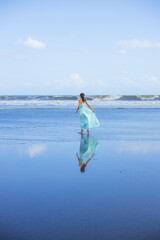 Young woman walking barefoot on empty beach. Full body portrait. Slim Caucasian woman wearing long dress. View from back. Water reflection. Waves on horizon. Vacation in Asia. Travel concept.