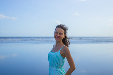 Fototapeta na wymiar Portrait of beautiful Caucasian woman on the beach. Summer vacation in Asia. Smiling young woman wearing dress. Sunny day. Blue sky. Ocean with horizon line. Travel lifestyle. Bali