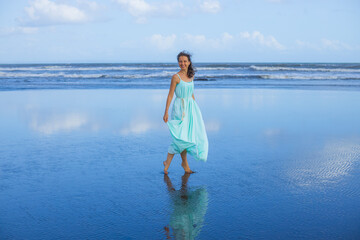 Happy smiling woman walking barefoot on empty beach. Full body portrait. Slim Caucasian woman wearing long dress. Water reflection. Blue sky. Summer vacation in Asia. Travel concept. Bali