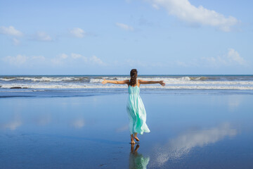 Young woman walking barefoot on empty beach. Full body portrait. Slim Caucasian woman wearing long dress. View from back. Water reflection. Freedom concept. Vacation in Asia. Bali