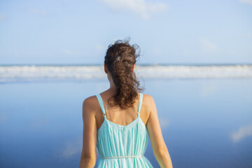 Fototapeta na wymiar Blurred back view portrait of beautiful Caucasian woman on the beach. Summer vacation in Asia. Young woman wearing dress. Blue sky. Ocean with horizon line. Travel lifestyle. Bali