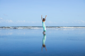 Slim woman walking barefoot on empty beach. Hands raised up. Caucasian woman wearing long dress. View from back. Water reflection. Blue sky. Vacation in Asia. Freedom concept. Bali
