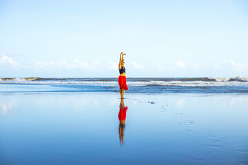 Young woman walking barefoot on the beach. Hands raised up. Caucasian woman wearing red skirt. Enjoy time on the beach. Water reflection. Summer vacation in Asia. Travel concept. Bali