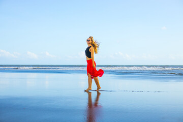Happy woman walking barefoot on the beach. Full body portrait. Caucasian woman wearing red skirt. Enjoy time on the beach. Water reflection. Summer vacation in Asia. Travel concept. Bali
