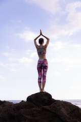 Woman standing on the rock, practicing yoga. Young woman raising arms with namaste mudra at the beach. Blue sky background. Yoga retreat in Bali. View from back. Copy space.