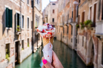 Close up of female hand holding strawberry gelato ice cream cone in front of canal with houses on...