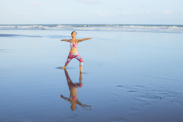 Beach yoga. Caucasian woman practicing Virabhadrasana II, Warrior II Pose. Strong body. Healthy lifestyle. Water reflection. Yoga retreat. Outdoor exercise. Copy space. Bali