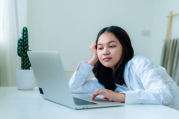 Portrait Cute Asian teen with long hair Wearing a white shirt, sitting online studying with a laptop on the bedroom table with a thoughtful face.