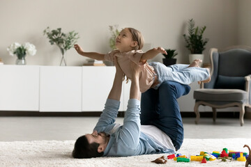 Caring young father lying on warm floor carpet, lifting little daughter pretending flying with...
