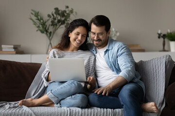 Happy young couple using laptop, relaxing on cozy couch at home together, smiling beautiful woman and man in glasses looking at computer screen, chatting online by video call or watching movie
