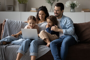 Happy parents with kids using laptop at home together, sitting on cozy couch, smiling mother and father with little son and daughter looking at computer screen, watching cartoons or movie, video call