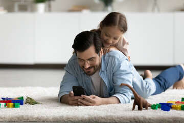 Fototapeta premium Smiling little daughter lying on father back, using smartphone together, having fun on warm floor at home, happy dad in glasses with girl kid looking at mobile device screen, watching cartoons