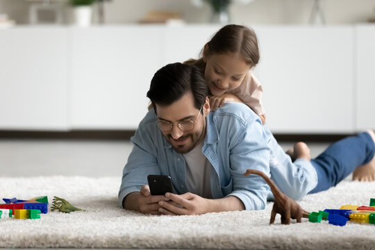 Smiling little daughter lying on father back, using smartphone together, having fun on warm floor at home, happy dad in glasses with girl kid looking at mobile device screen, watching cartoons