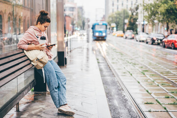 Woman waiting for public transport on bus stop looking to the smartphone, using application for...