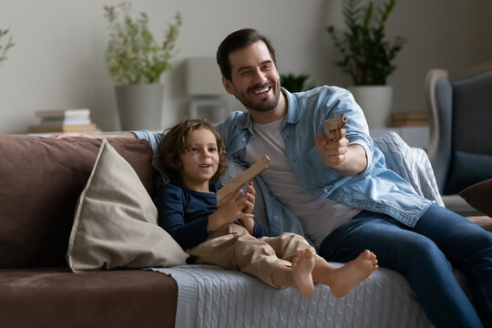 Happy father and little son having fun with wooden toy guns, sitting on comfortable couch at home, smiling dad with adorable 5s boy child engaged in funny activity, spending leisure time together