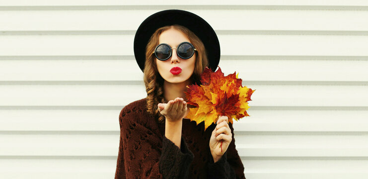 Portrait of beautiful young woman with yellow maple leaves blowing her lips sending sweet air kiss wearing a brown knitted poncho, black round hat on white background