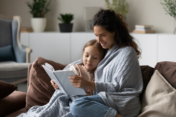 Smiling caring mother with little daughter reading book together, happy mom with adorable girl child wrapped in warm blanket sitting relaxing on comfortable couch, engaged in educational activity - obrazy, fototapety, plakaty